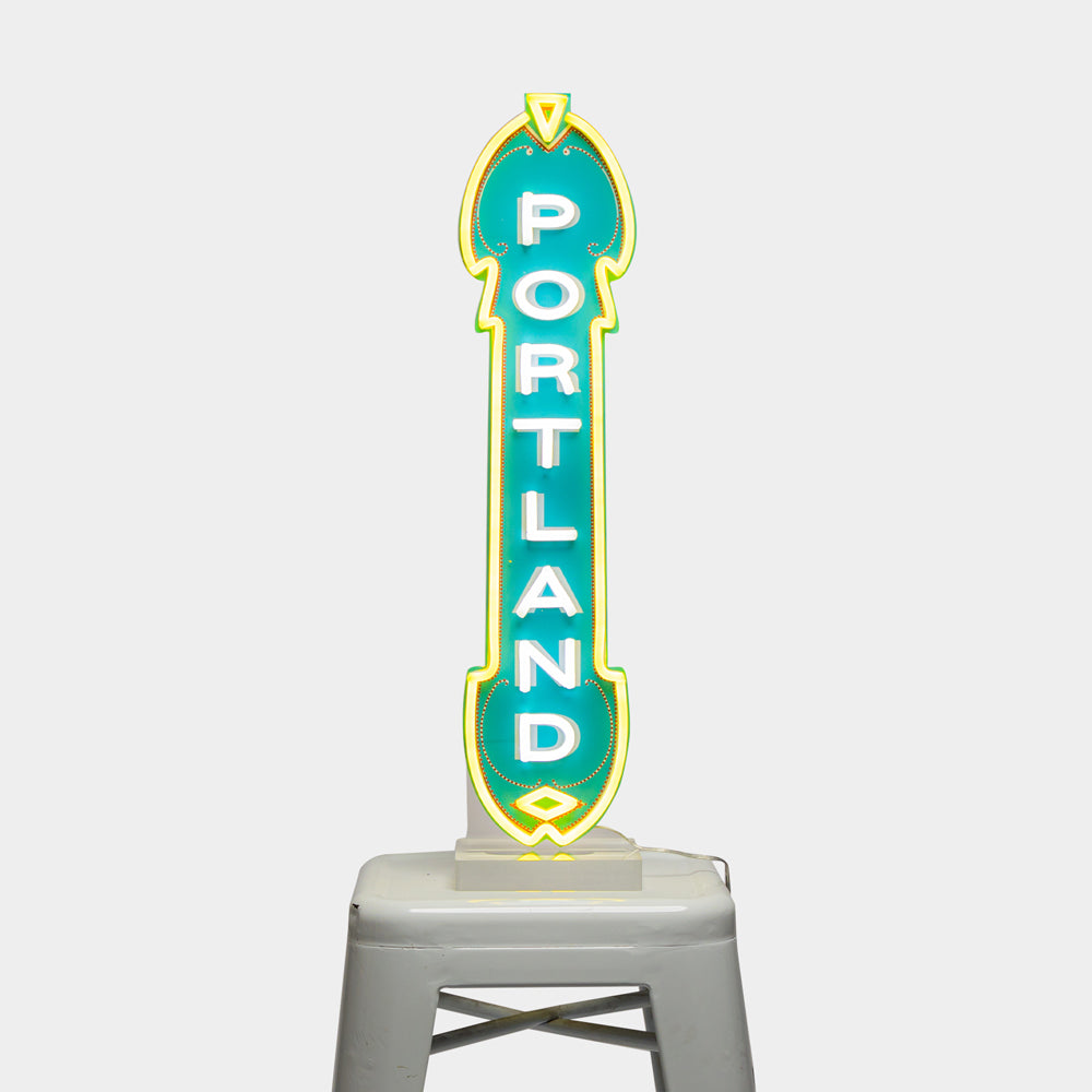 The LED Marquee Sign showcases the word "Portland" in bold, white letters set against a green background and outlined with yellow. This energy-efficient sign is mounted on a minimalist gray stool, making it pop against the plain white backdrop.