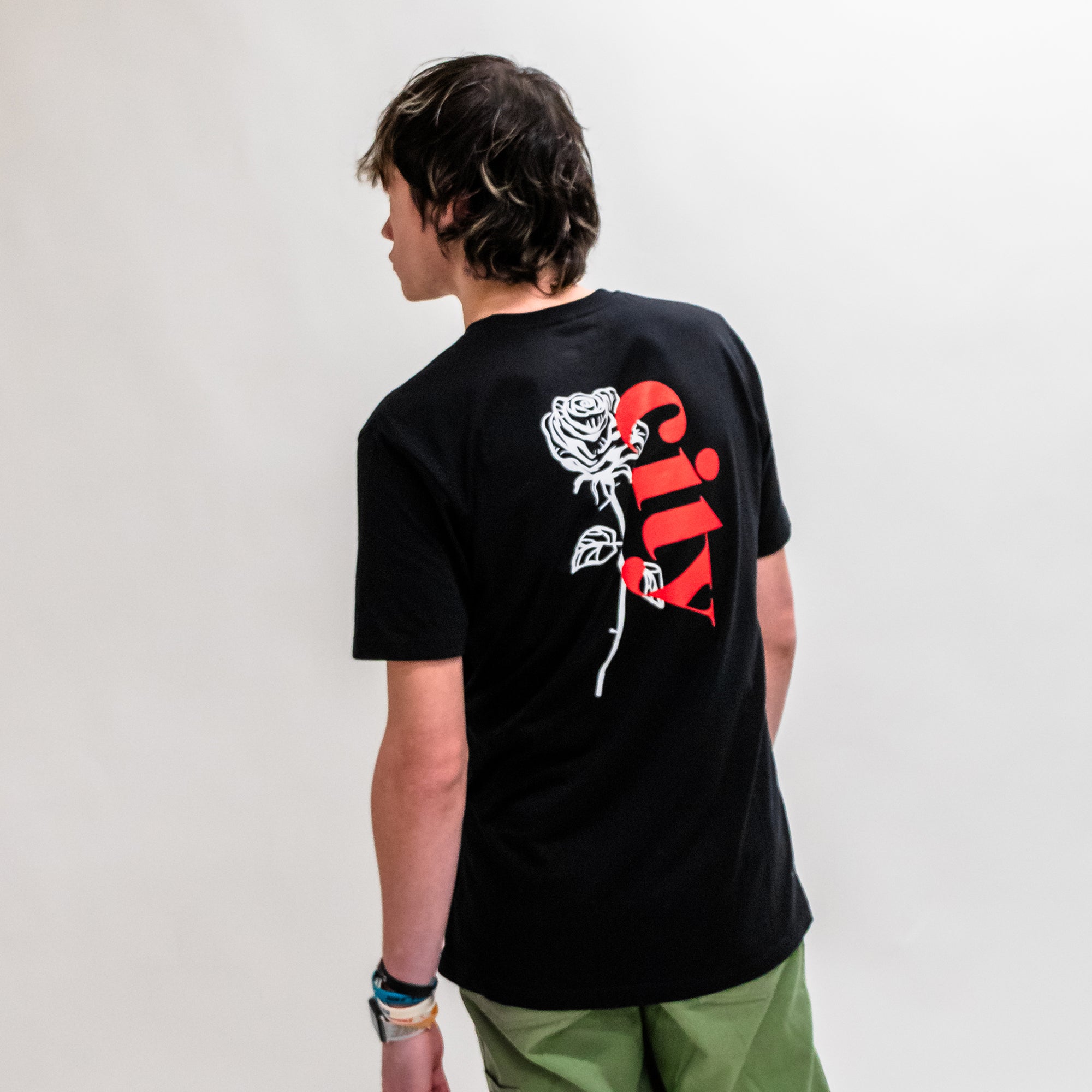A person facing away, wearing a black Soft-Blend Rose City "P" Tee featuring a graphic of a white rose and the word "Silly" in red on the back. The individual has shoulder-length hair and is also wearing green pants. The background is a plain light gray.