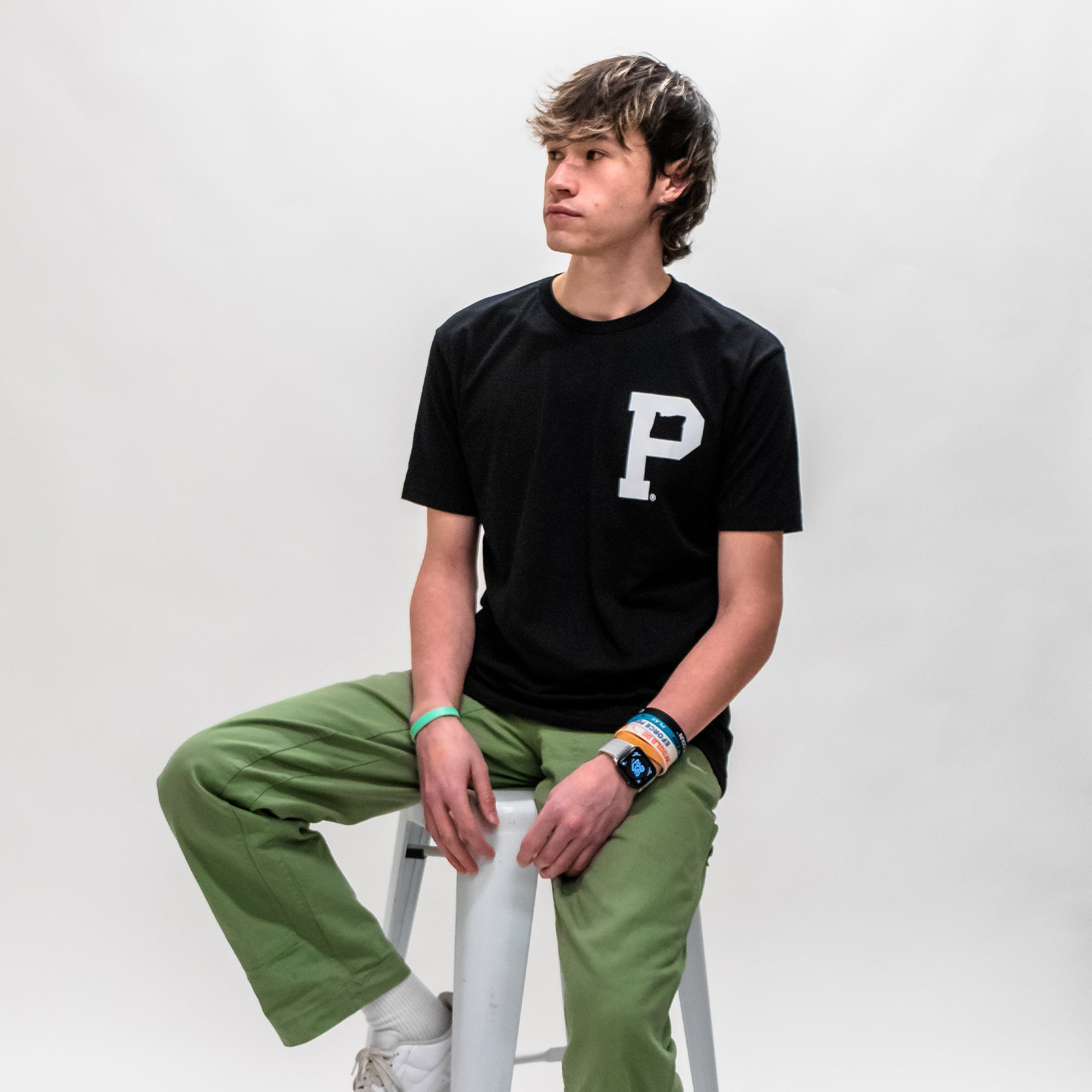 A young man, wearing a black Soft-Blend Rose City "P" Tee featuring a large "P" on the chest and green pants, is seated on a white stool against a plain, light background. He gazes off to the side with a neutral expression, his left wrist adorned with a watch and several wristbands.