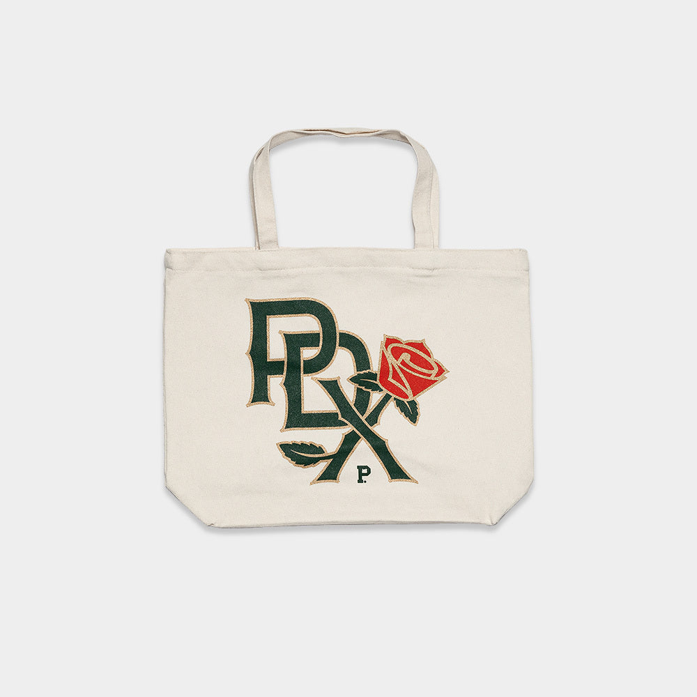 The PDX Rose Zip Tote is a beige canvas bag with two sturdy handles. It features the letters "PRLX" in dark green and a red rose in an artistic design, prominently showcased at the center of the tote. Additionally, it has a durable zipper top for added security.