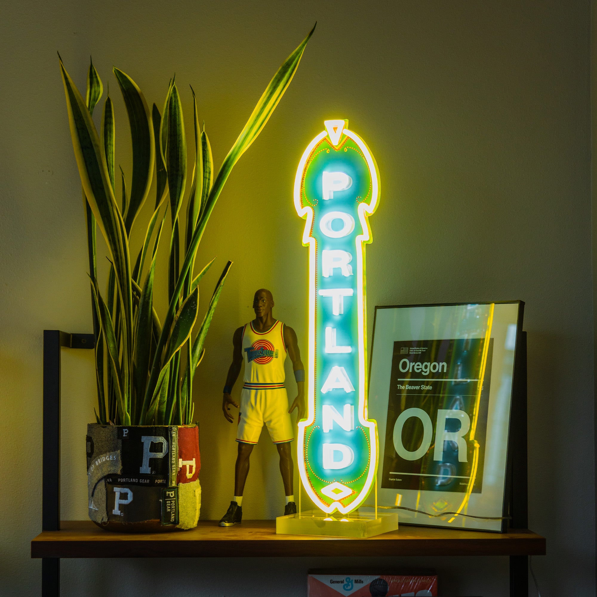 A shelf featuring a potted plant, a basketball player figurine, an energy-efficient yellow LED Marquee Sign that reads "Portland," and a framed poster of Oregon creates a vibrant, eclectic display. The neon sign and the plant work together to stand out against the dimly lit background wall, highlighting the bright elements on the shelf.