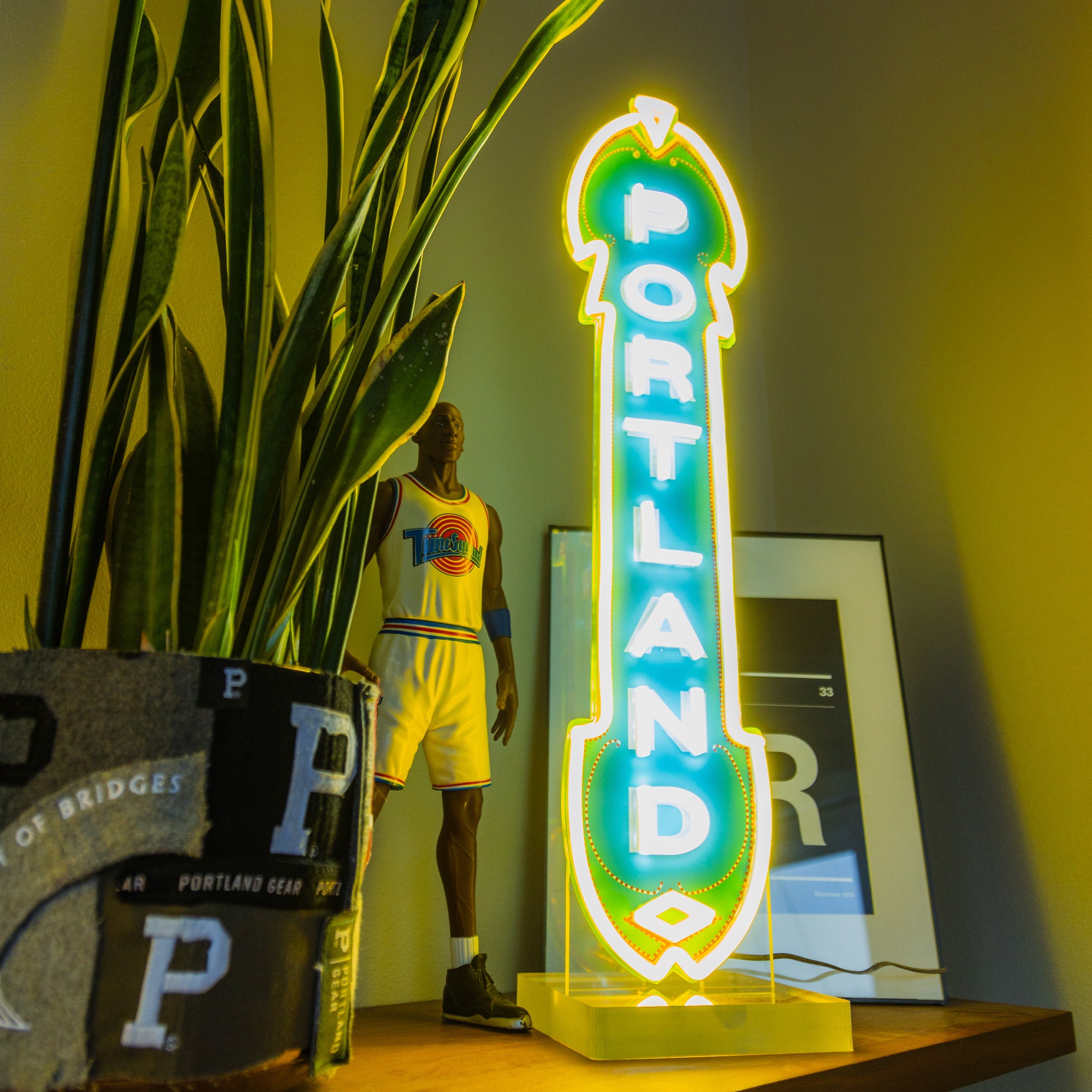 A neon sign with the word "PORTLAND" glows in vibrant blue and yellow, resembling an LED Marquee Sign known for its energy efficiency. Beside it sits a potted plant featuring a "P" design on the container. In the background, there is a figurine of a basketball player dressed in a white jersey, with part of a framed piece of artwork visible behind the sign.