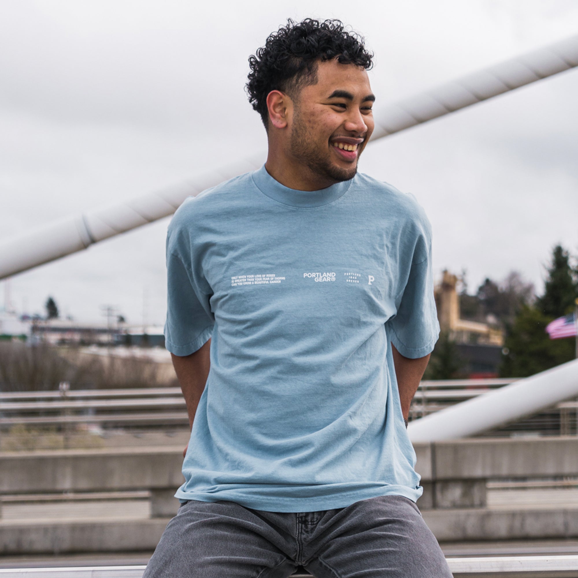 A man with short curly hair and a beard is smiling while sitting on a bridge in Rose City. He is wearing the All-American Slogan Tee - Sky, a light blue T-shirt made from 100% cotton with white text, along with grey pants. The background features a cloudy sky and blurred greenery.