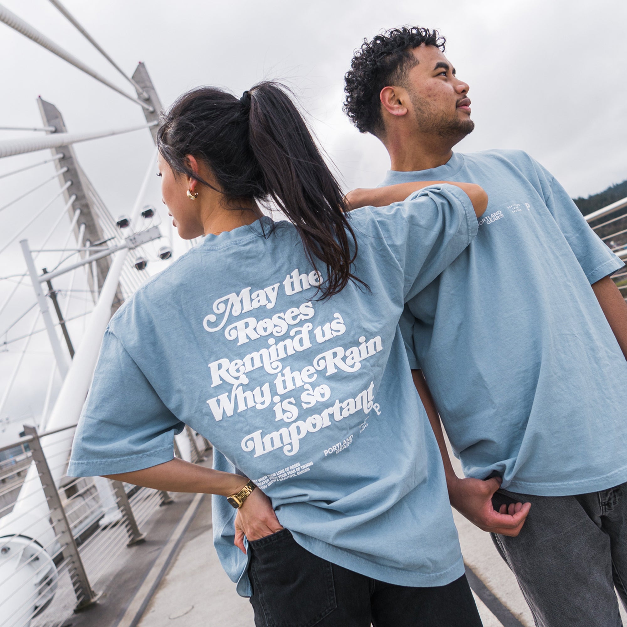 Two people stand side by side outdoors, wearing All-American Slogan Tees in the Sky color. The T-shirts are made of 100% cotton and feature a light blue oversized design. The woman faces away from the camera, displaying text that says, "May the Roses Remind us Why the Rain is so Important." Both individuals gaze toward Rose City.