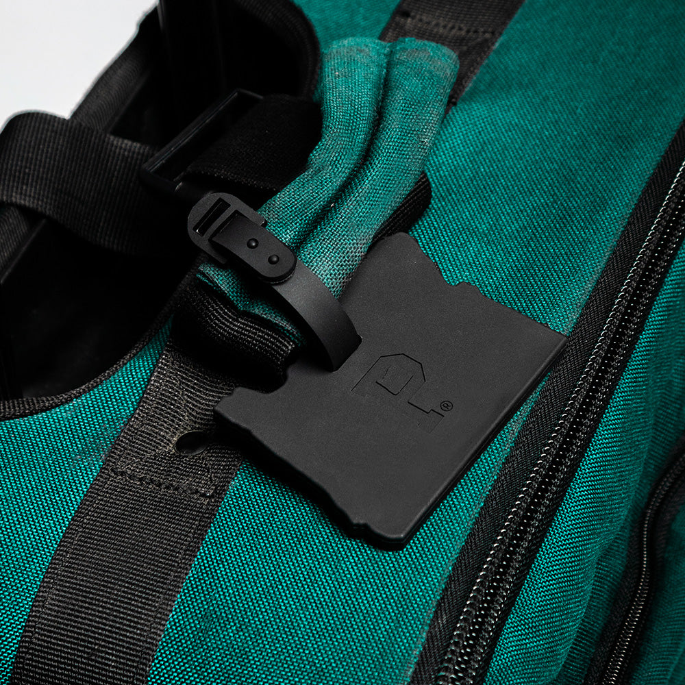 A close-up of a green fabric bag with black zippers and straps. Attached to one of the secure attachment straps is a Cascade Luggage Tag in black, uniquely shaped like the state of Washington, featuring an outline of a house and a registered trademark symbol.