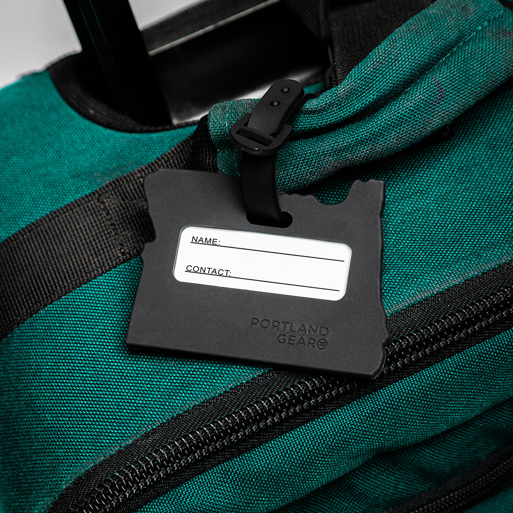 Close-up of a black **Cascade Luggage Tag** shaped like the state of Oregon, securely fastened to a green bag with black zippers. The silicone luggage tag features spaces for "Name" and "Contact" information with the brand text "Portland Gear" underneath, though no details are currently filled in.