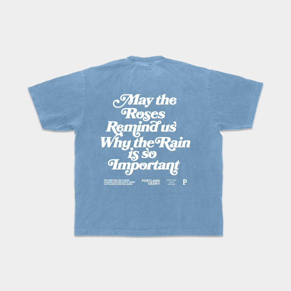 The All-American Slogan Tee - Sky is a light blue, 100% cotton t-shirt with white text on the back that reads "May the Roses Remind us Why the Rain is so Important." Smaller text beneath this slogan mentions "Portland Gear" along with other intricate details. Show your Rose City pride with this stylish tee, which comes in sustainable packaging. The shirt is showcased against a plain background.