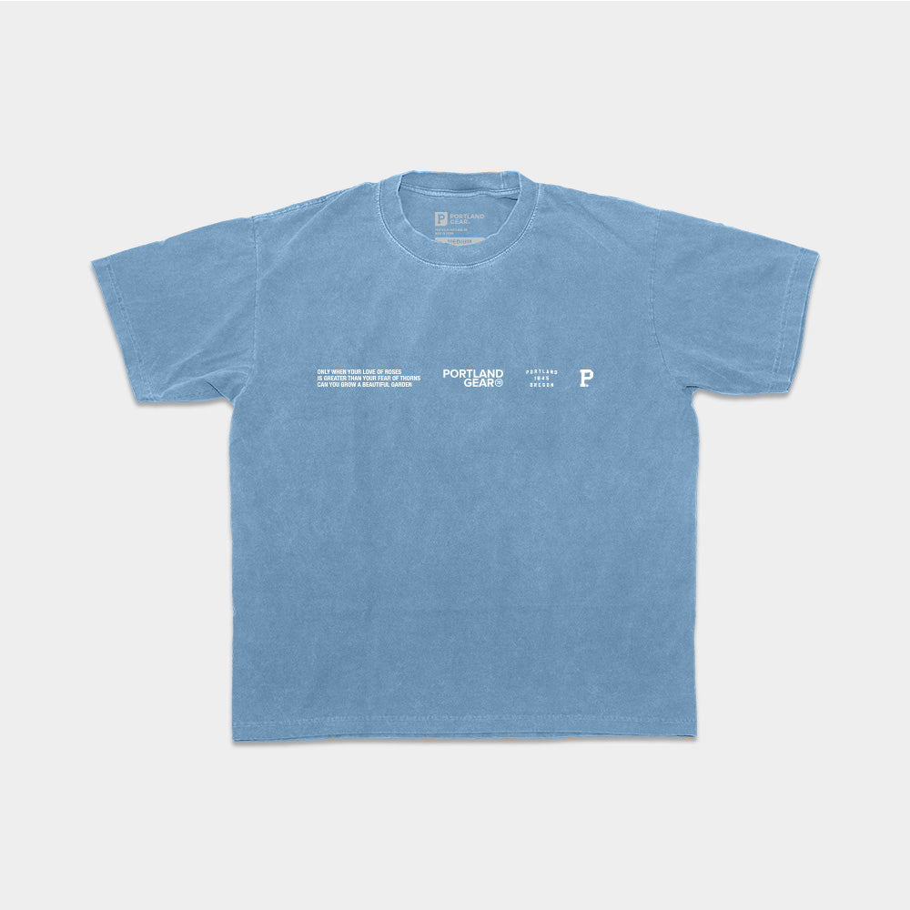 The All-American Slogan Tee - Sky is a light blue T-shirt featuring the text "Portland Gear" and a small illustration of a map of Portland on the front. Made from 100% cotton, this shirt also displays a poem and logos near the chest. Its simple, casual design with short sleeves and a crew neck perfectly captures the essence of Rose City.