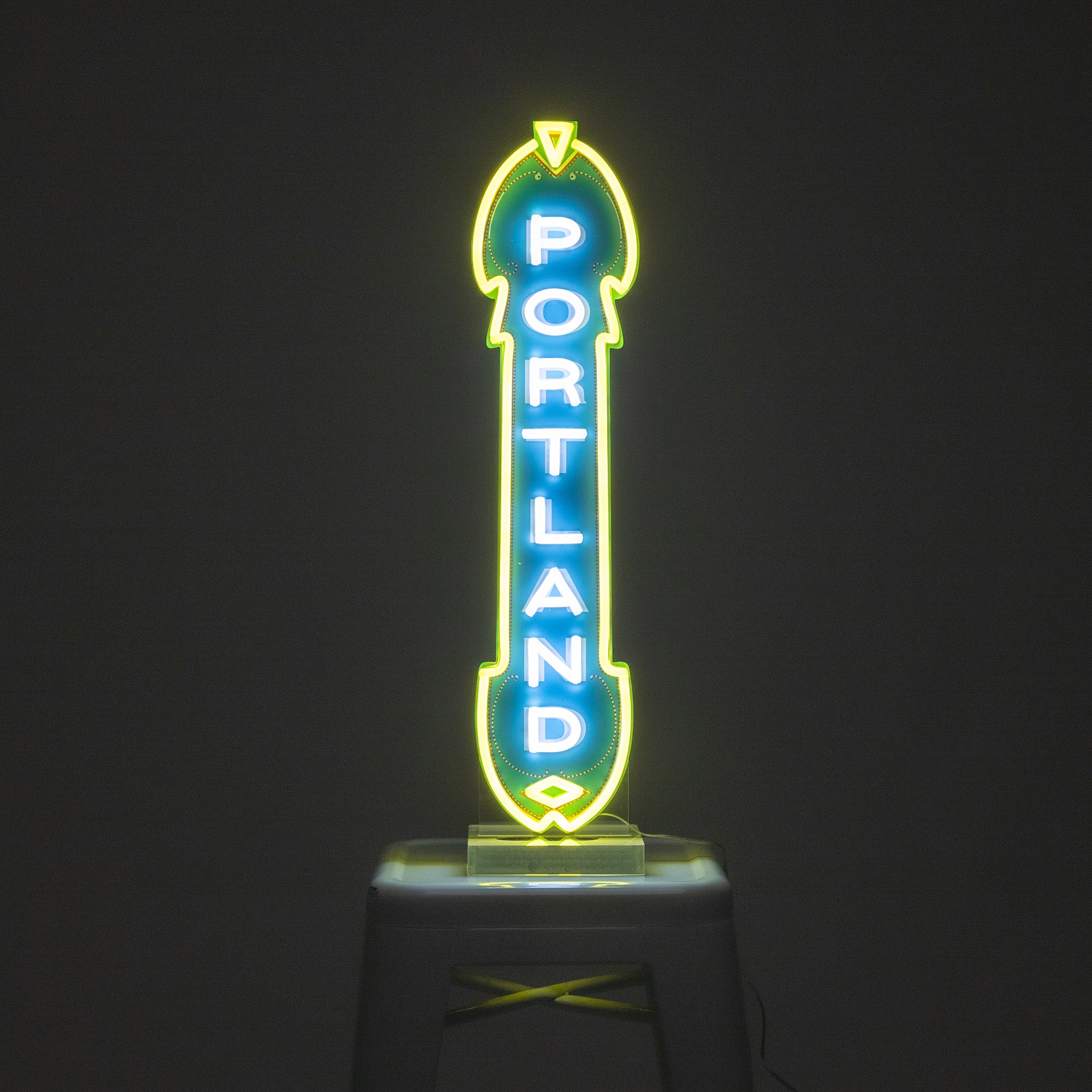 A custom LED Marquee Sign in the shape of an arrow, glowing in green, blue, and yellow, with the word "PORTLAND" written vertically in the center. This energy-efficient sign is displayed against a dark background and placed on a stool.