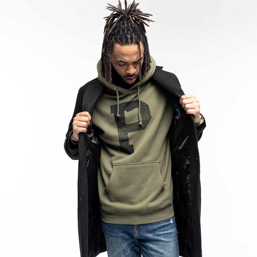 A man with dreadlocks is wearing a Soft-Blend "P" Hoodie in olive, featuring a large black "P" on the front. He is also wearing a black coat, which he is holding open with both hands. He is looking down and standing against a plain white background.