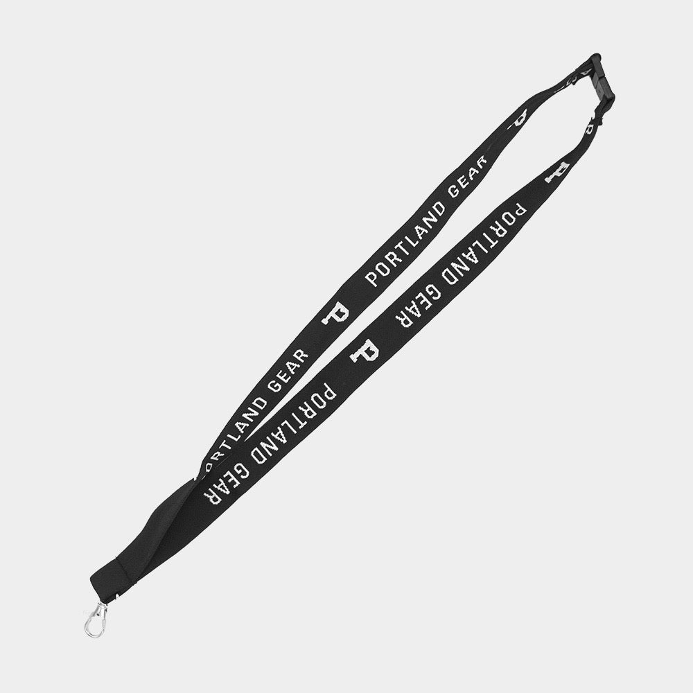 A black Lanyard from Portland Gear features white "PORTLAND GEAR" text repeated along its length in a double-sided woven design. It includes a metal clip on one end and a plastic buckle closer to the top. The background is white.