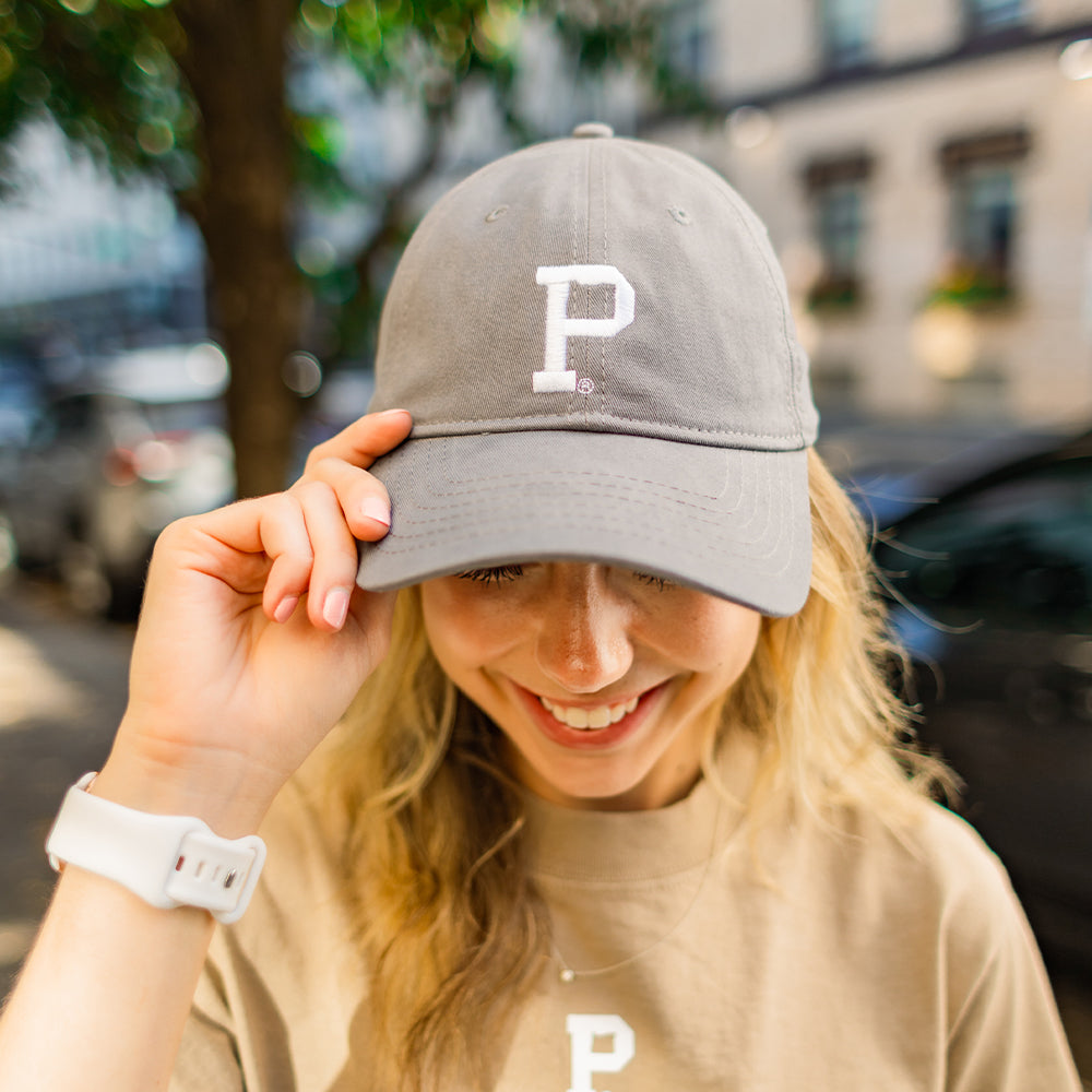 A smiling person with blonde hair wears the "Dad Hat - Dark Grey," featuring a white "P" on it, which represents a popular Shell product. They are also dressed in a beige t-shirt with the same "P" logo on the front. The person is outdoors, holding the brim of their hat with one hand, while a blurred street with trees and buildings appears in the background.