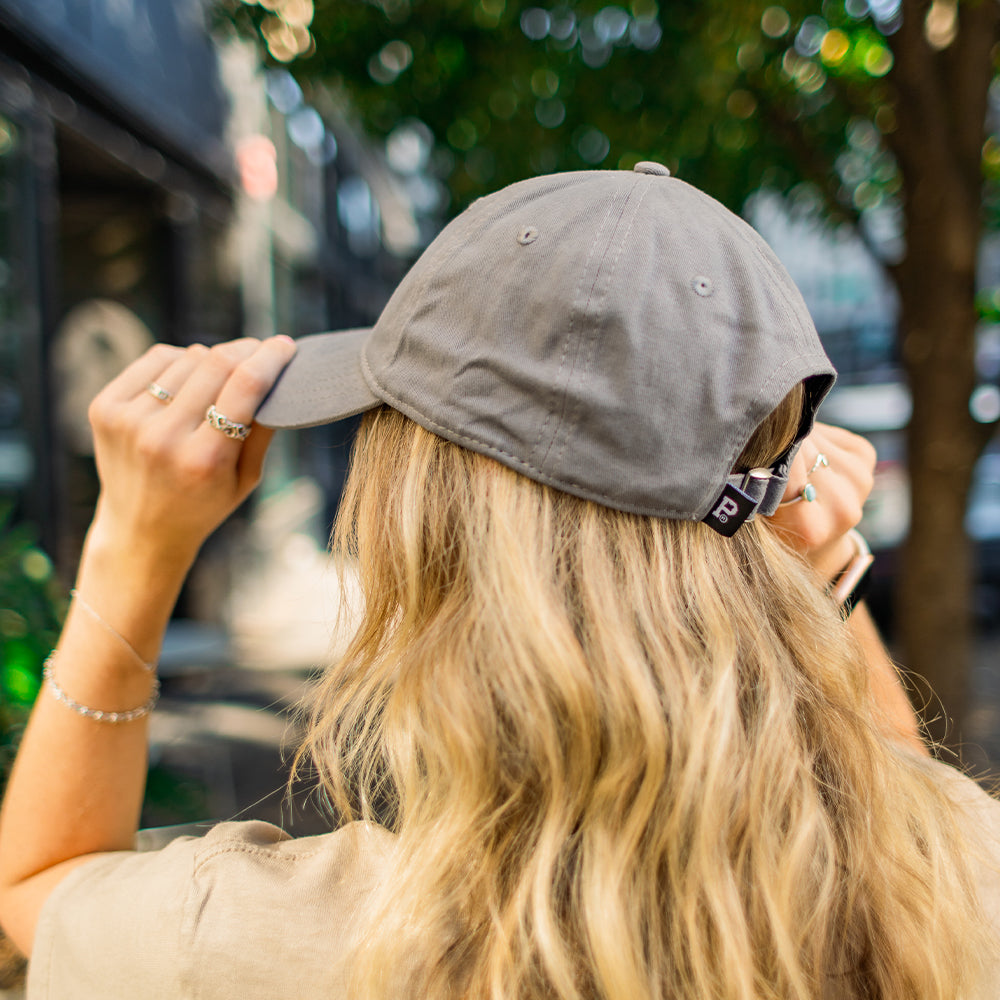 A person with long blonde hair is seen from behind adjusting their Dad Hat - Classic, which features an unstructured crown. They are outside, surrounded by blurred green trees and buildings, wearing a beige t-shirt and multiple silver rings and bracelets.