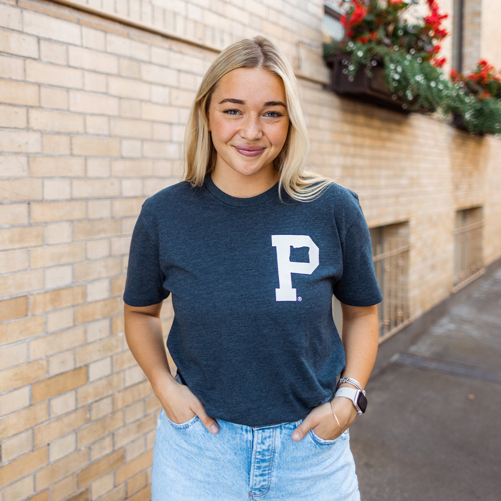 A person with long blonde hair stands smiling, hands in pockets, wearing a heather navy Soft-Blend "P" Tee - Classic and light blue denim shorts. They are in an outdoor setting with a brick wall and flower boxes in the background.