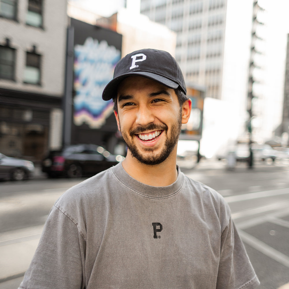 A smiling man with a beard and mustache is wearing a Dad Hat - Classic, which features an unstructured crown and the letter "P" on it, along with a gray cotton t-shirt also featuring the letter "P." He is standing on a city street with blurred buildings and cars in the background.