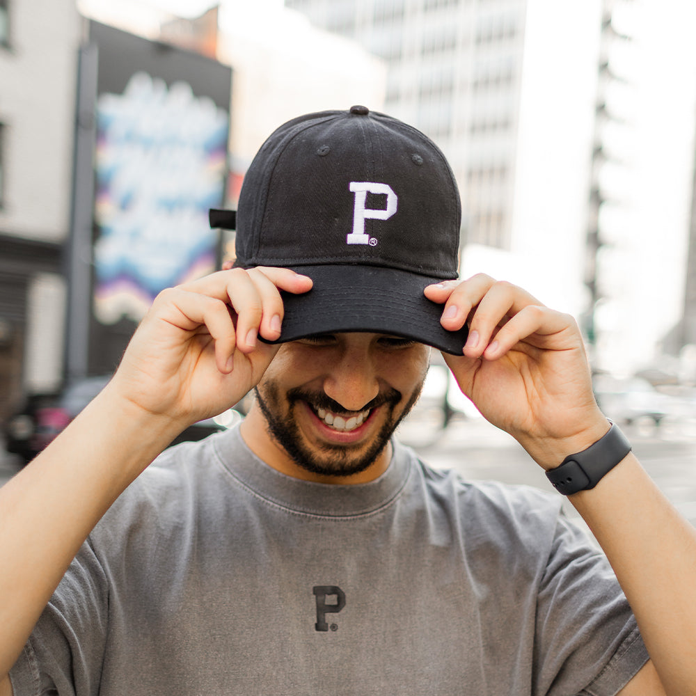 A man wearing a gray t-shirt and a Dad Hat - Black with the letter "P" stands on a city street. Adjusting the brim of his hat, he smiles warmly. Urban buildings and a colorful mural form the backdrop of this vibrant urban setting, showcasing the city as a lively shell of creativity.
