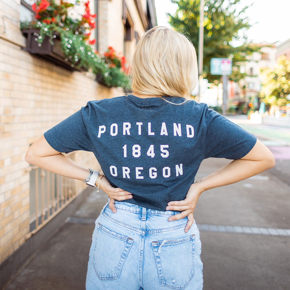 A person with long blonde hair stands with their back to the camera, wearing a dark blue Soft-Blend "P" Tee - Classic from Portland Gear that reads "Portland 1845 Oregon" and light blue jeans. The sidewalk is adorned with buildings, flowers, and greenery in the background.
