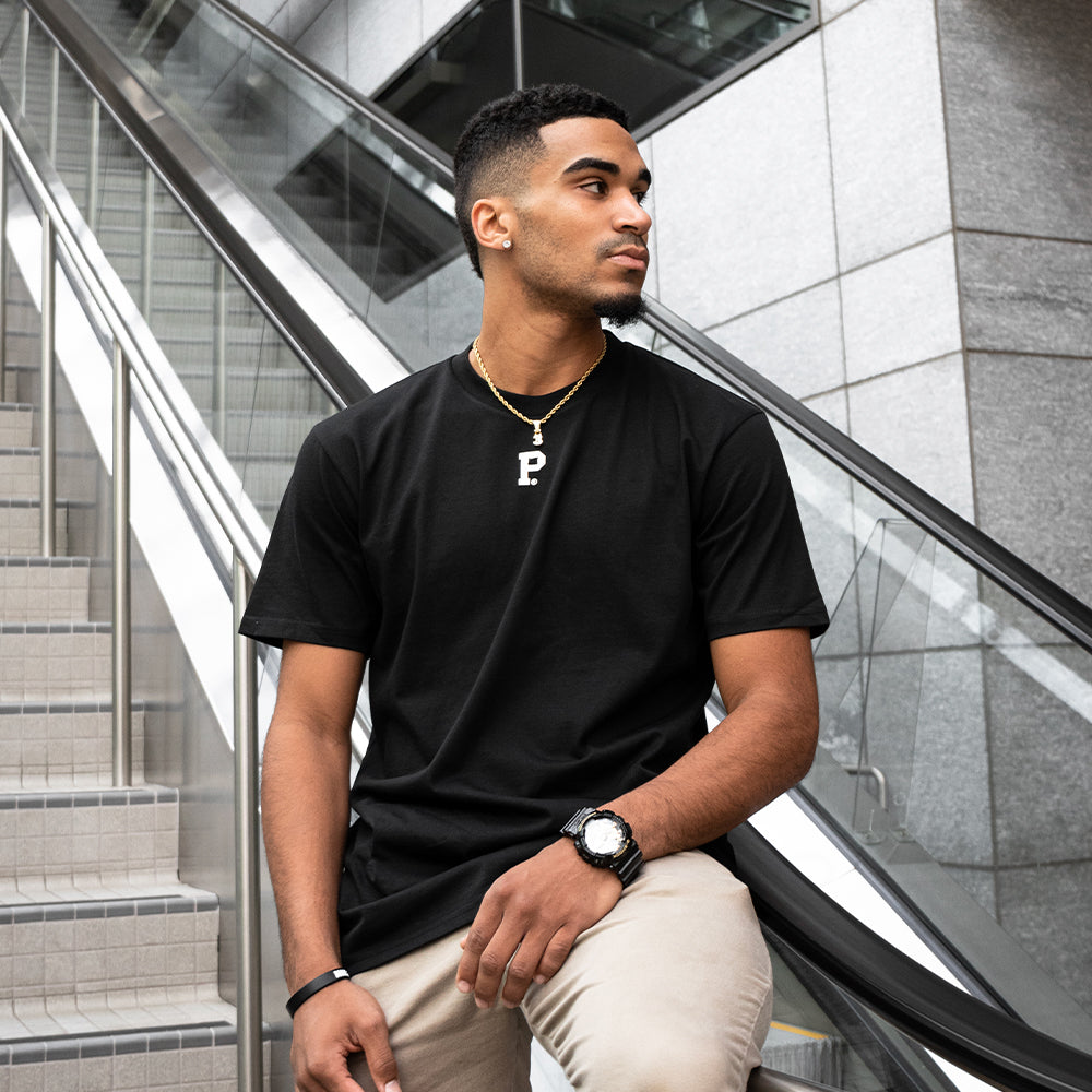A man wearing a HeavyKnit Centerpiece Tee - Black with a large "P" pendant necklace and a wristwatch sits on a stairway railing next to an escalator, looking to his left. The background features modern architectural elements with glass and concrete. His attire exudes quality with its high-density screenprint design.