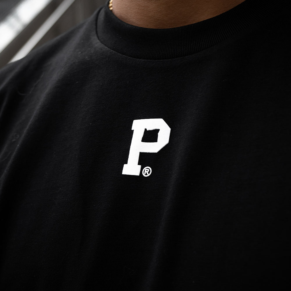 Close-up of a person wearing the HeavyKnit Centerpiece Tee - Black, featuring a white letter "P" and a registered trademark symbol on the chest. The shirt boasts high density screenprint on heavyweight cotton. The background is slightly blurred, emphasizing the shirt design.