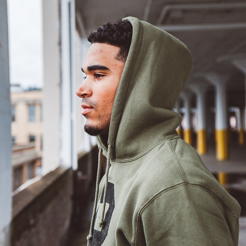 A man wearing the Soft-Blend "P" Hoodie in olive stands in an industrial-looking building, gazing out of a window. The background features large columns and a partially blurred view of buildings outside. The mood appears contemplative.