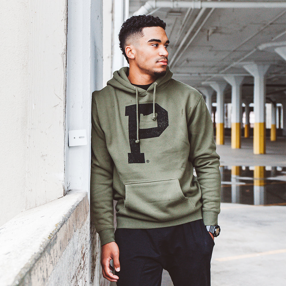 A person wearing an olive Soft-Blend "P" Hoodie with a large black "P" on the front, black pants, and a watch stands in a spacious, industrial-looking indoor area. They are leaning against a wall, looking away from the camera.