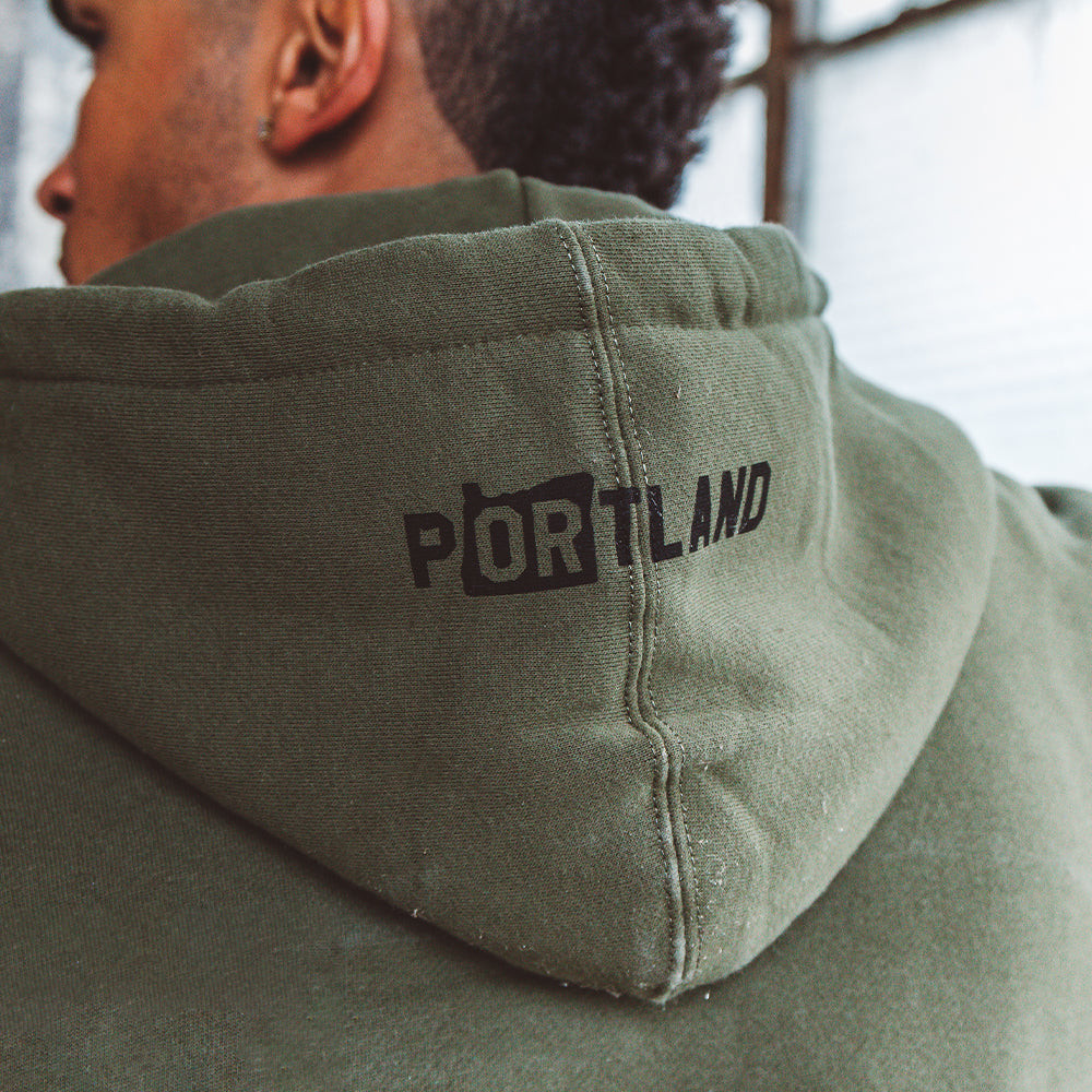 A person wearing the Soft-Blend "P" Hoodie in olive with "PORTLAND" printed in black on the back of the hood. The focus is on the text, while the person's face is turned away from the camera. The background is slightly out of focus.