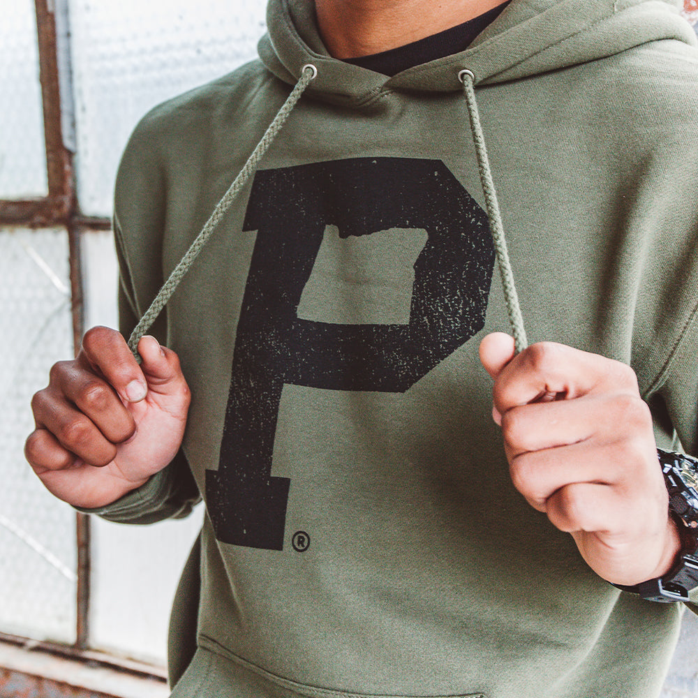 A person wearing the Soft-Blend "P" Hoodie in olive, which features a prominent letter "P" on the front, holds the drawstrings. Their face is out of view, and the background includes a frosted glass window alongside part of a brick wall.