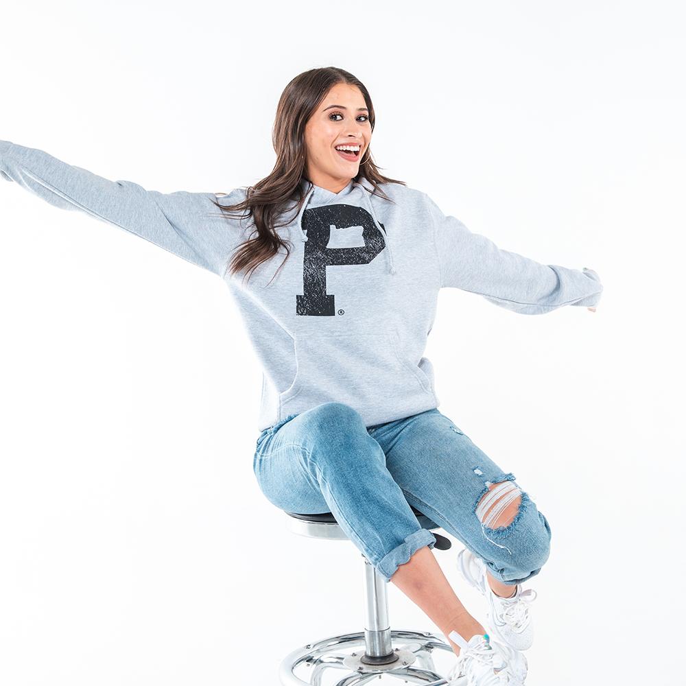 A woman in a Soft-Blend "P" Hoodie - Grey with a large black letter "P" on the front, blue ripped jeans, and white sneakers is sitting on a stool with her arms outstretched, smiling widely against a plain white background.