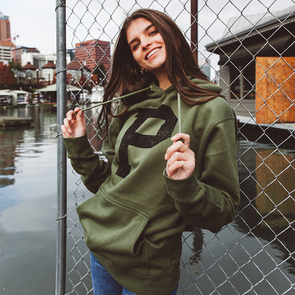 A smiling woman wearing a Soft-Blend "P" Hoodie in olive green, featuring a large black "P" on the front, stands near a chain-link fence by the water. She is holding the hood strings with both hands. Buildings and boats are visible in the background.