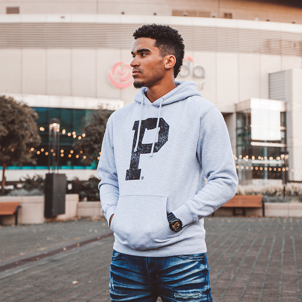 A person stands confidently outdoors wearing a Soft-Blend "P" Hoodie - Grey, an athletic sweatshirt featuring a prominent black "P" on the front. They have short, curly hair and are also dressed in blue distressed jeans. In the background, there is a building with a circular architectural design and string lights.