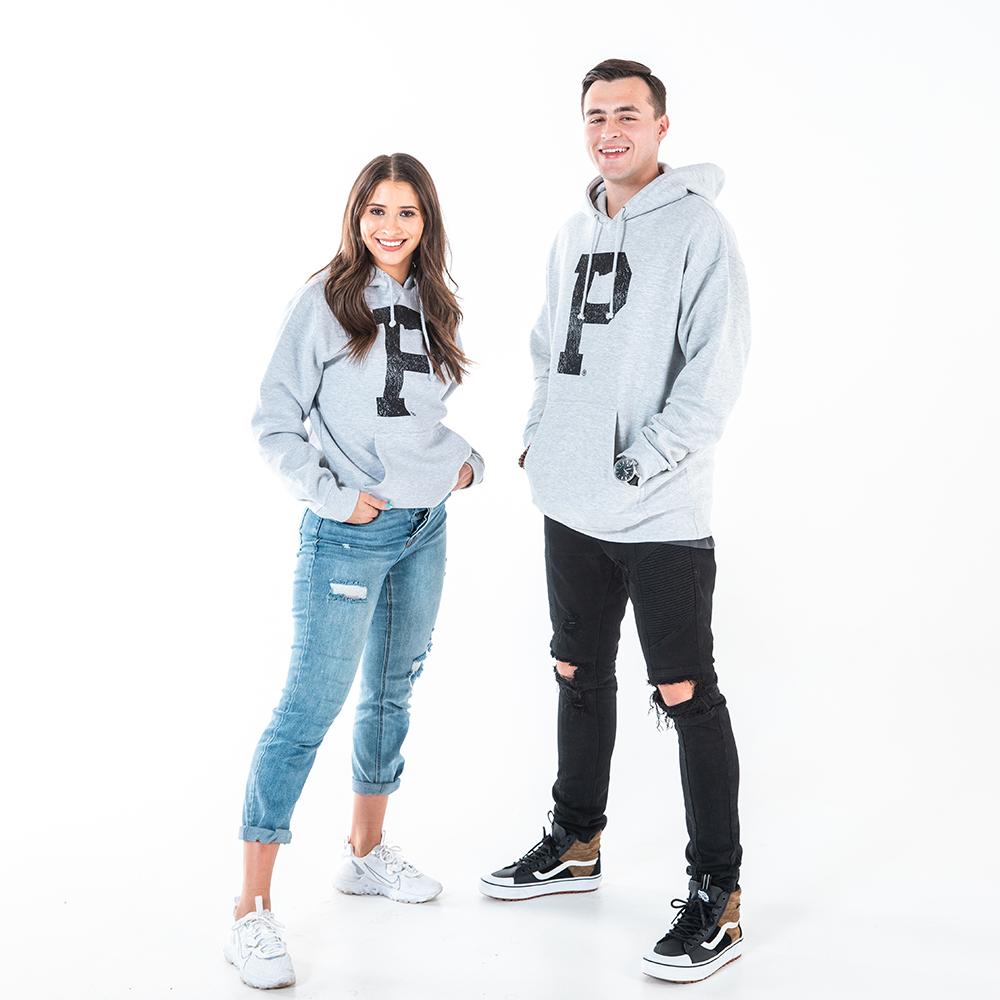 A smiling woman and man stand against a plain background, both wearing the Soft-Blend "P" Hoodie in grey. The woman, in rolled-up jeans and sneakers, stands to the left. The man, in ripped black jeans, sneakers, and a watch, stands to the right.