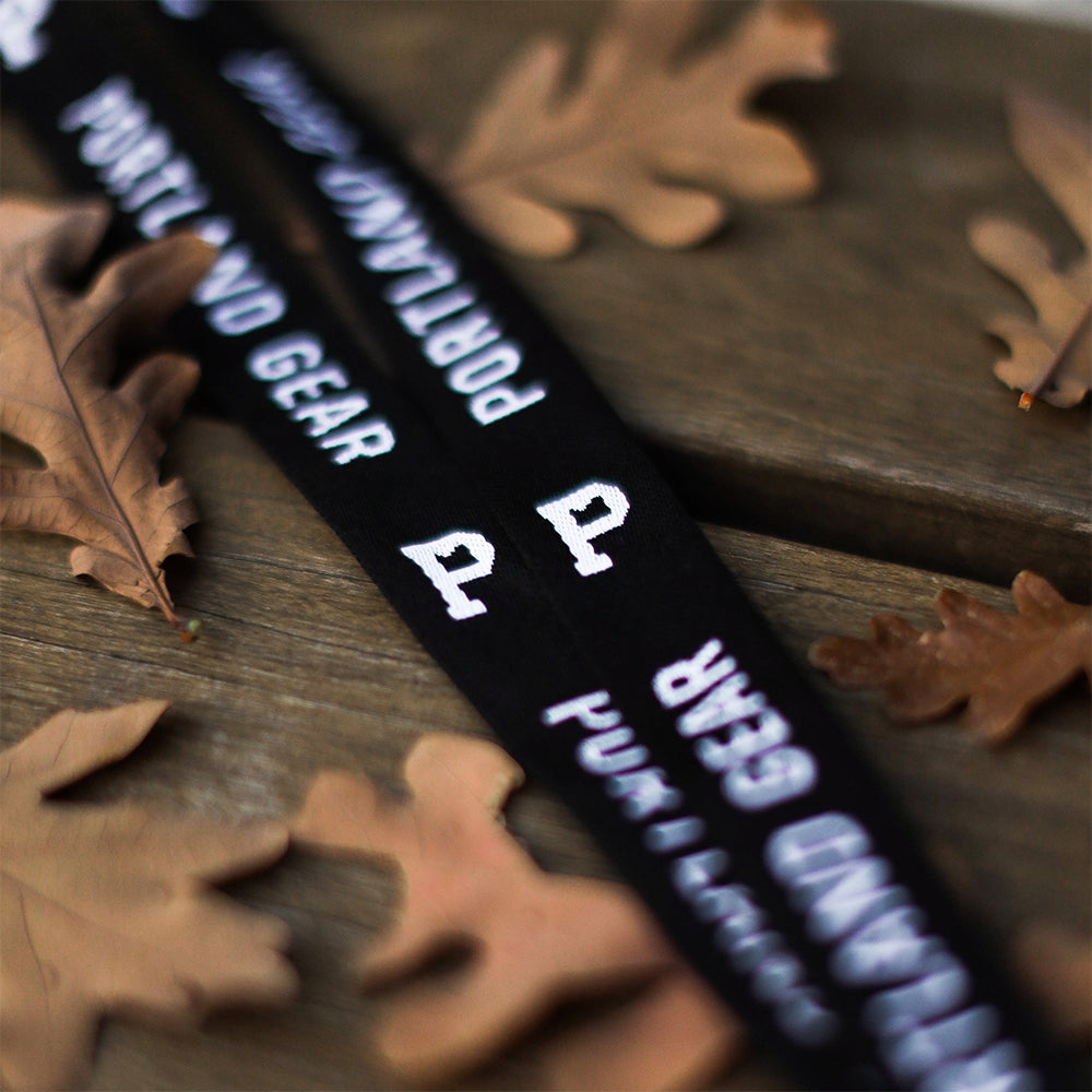 A close-up of black lanyards from Portland Gear, showcasing white text and the "P" logo with a double-sided woven design. The lanyards are spread out on a wooden surface, adorned with dry autumn leaves.
