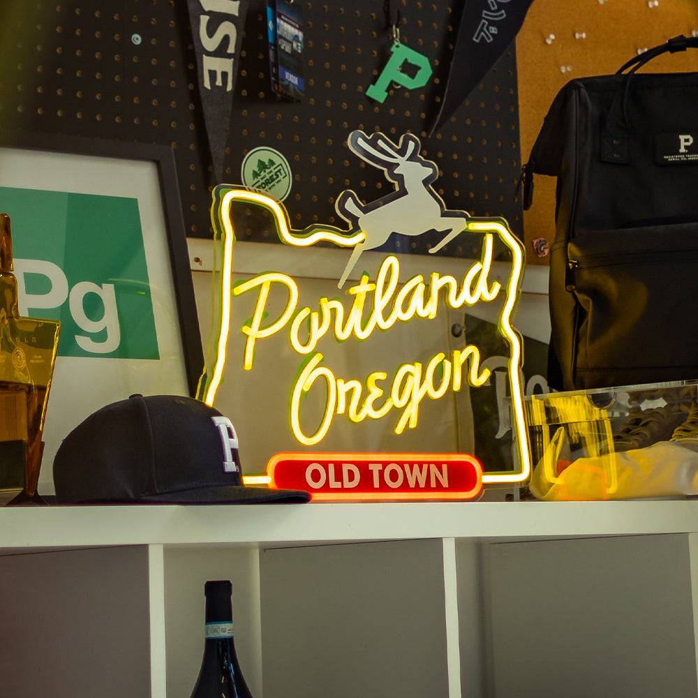 A brightly lit, energy-efficient LED Portland Sign is shaped like the state of Oregon and features the text "Portland Oregon" with an image of a leaping deer on top and "OLD TOWN" beneath. This custom LED sign sits on a white shelf next to various items, including a black cap, a framed poster, and a black backpack.