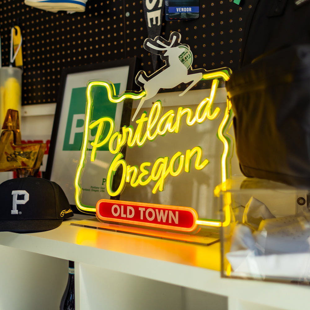 The LED Portland Sign, custom-made in the shape of the state of Oregon and featuring the words "Portland Oregon Old Town" with a leaping stag on top, is an energy-efficient decoration displayed on a shelf alongside various items such as a framed picture and a baseball cap with the letter "P".