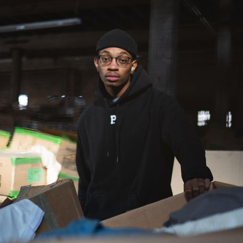 A person wearing a high-density white P logo black beanie and a HeavyKnit Centerpiece Hoodie - Black stands in a dimly lit warehouse. They are looking at the camera while leaning on a box filled with various items, including clothing and sheets. The background is filled with other large boxes and industrial equipment.