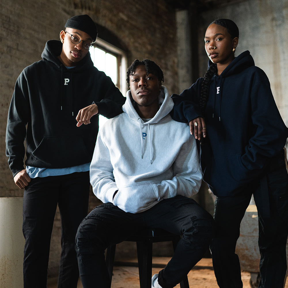 Three people pose confidently in a dimly lit space wearing hoodies. The person on the left wears a HeavyKnit Centerpiece Hoodie - Black, glasses, and a beanie; the person in the middle, seated, wears a light gray hoodie; and the person on the right sports a black hoodie with braids.
