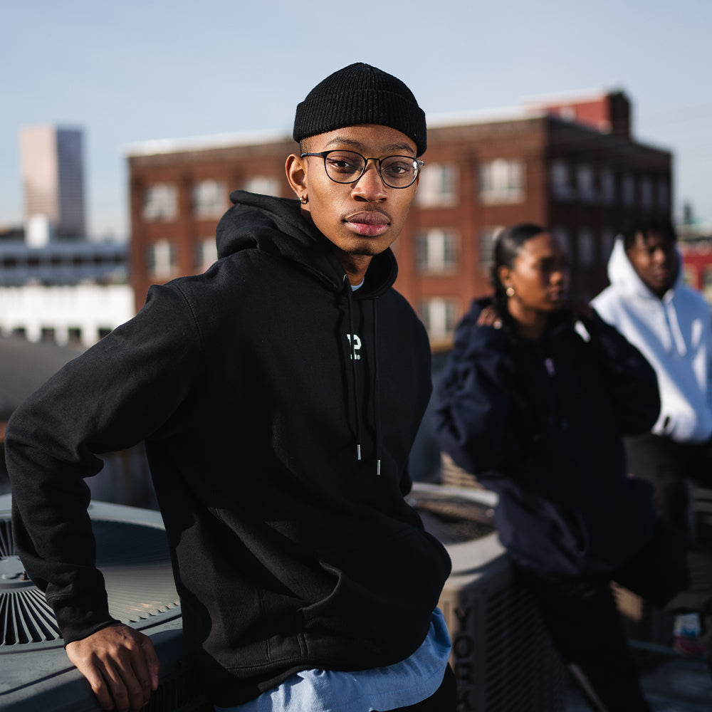 A young man wearing a black beanie, glasses, and a HeavyKnit Centerpiece Hoodie - Black, which features a high-density white P logo, stands confidently on a rooftop with two friends blurred in the background. Buildings and the cityscape are visible behind them. The mood is casual and urban.