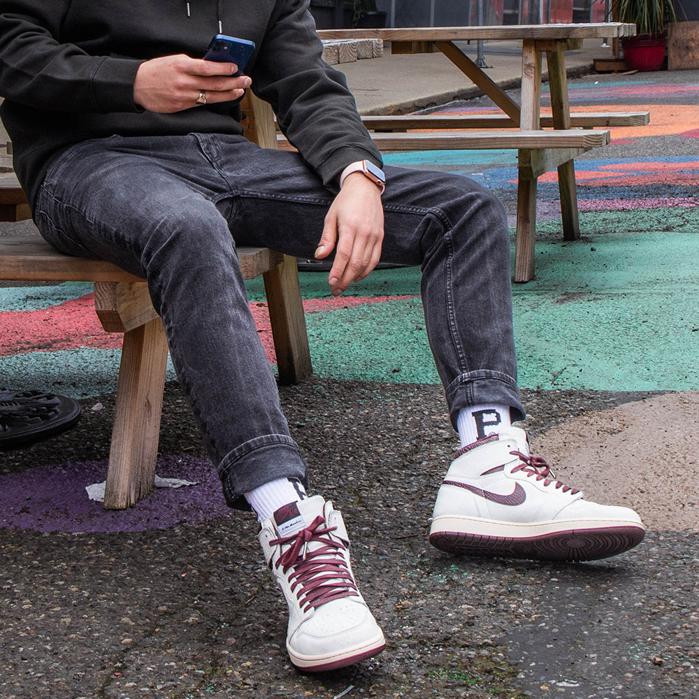 A person in a black hoodie and dark jeans sits on a wooden bench, holding a smartphone in one hand. They sport white Core Socks from Portland Gear featuring the iconic "P" logo, paired with white high-top sneakers with red laces. The ground beneath them features colorful painted patterns.
