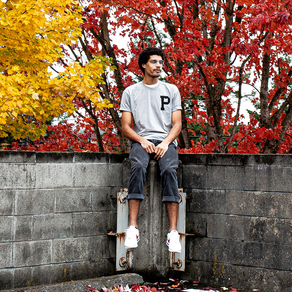 A man sits on a concrete wall against the vibrant fall backdrop, with trees displaying bright red and yellow leaves. He is wearing the Soft-Blend Rose City "P" Tee in grey, dark pants, and white sneakers, gazing into the distance.