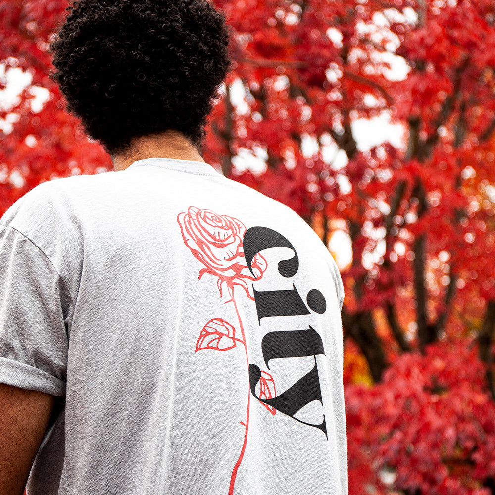 Person with curly hair wearing a Soft-Blend Rose City "P" Tee in light gray, standing in front of a tree with vibrant red leaves.