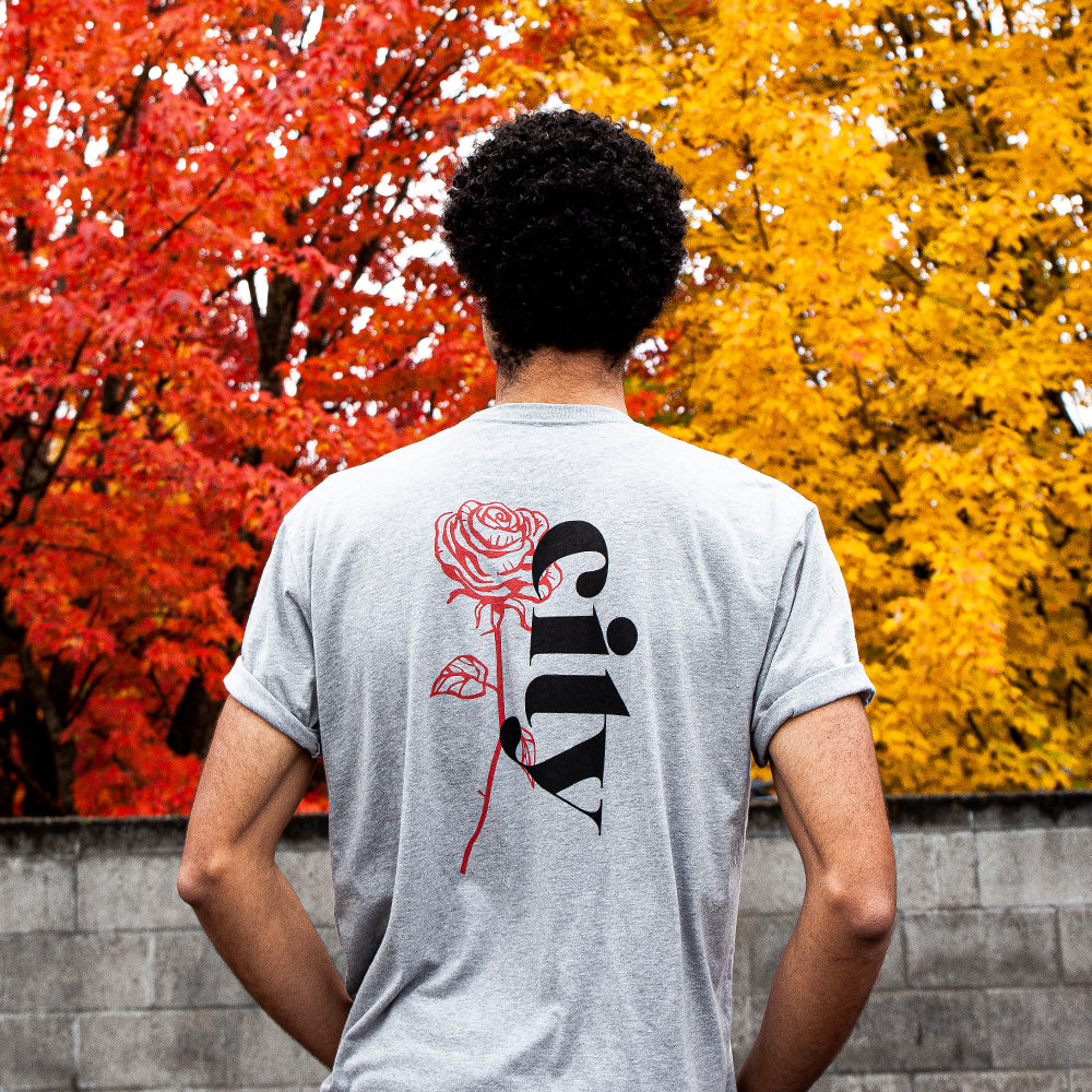 A person stands outdoors, facing away from the camera, wearing the Soft-Blend Rose City "P" Tee in grey. The light gray unisex cotton polyester tee features a rose and the word "city" on the back. Vibrant autumn foliage with prominently displayed red and yellow leaves creates a striking background. Sustainability is enhanced by eco-friendly packaging.