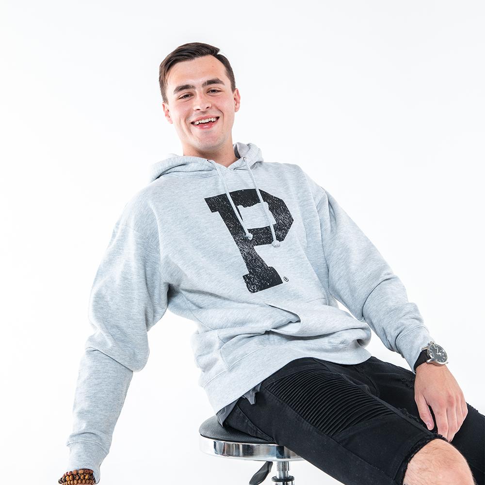 A young man with short dark hair is sitting on a stool against a plain white background. He is wearing an athletic grey Soft-Blend "P" Hoodie featuring a large black letter "P" on it, paired with black shorts. On his left wrist, he sports a watch, and on his right wrist, he wears a beaded bracelet. With a cheerful smile, he looks directly at the camera.