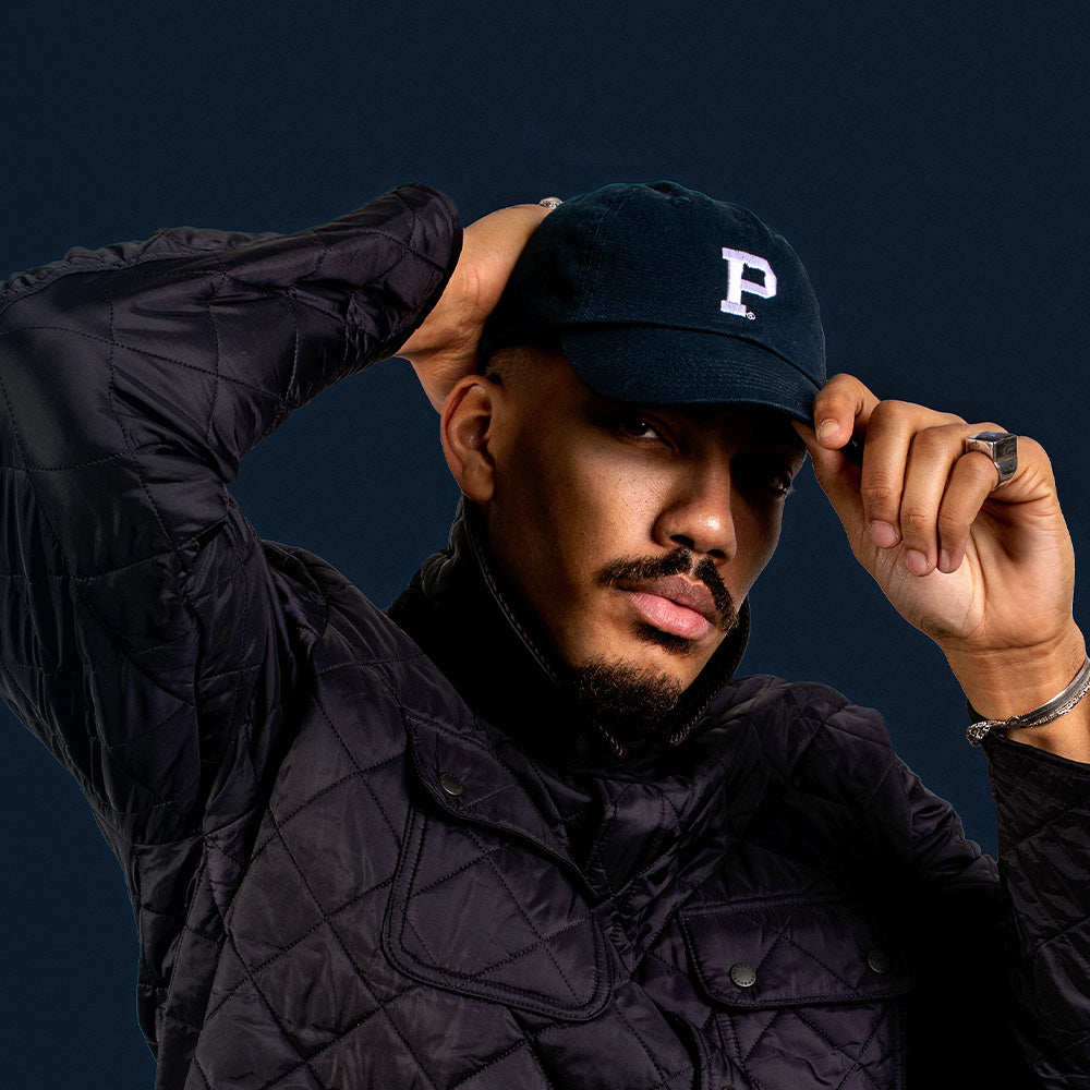 A person wearing a dark quilted jacket and a navy baseball cap featuring the letter "P" adjusts their cap with their right hand while resting their left arm on their head. The background is dark, creating a dramatic contrast with the person's attire, highlighting the quality of this Dad Hat - Navy.
