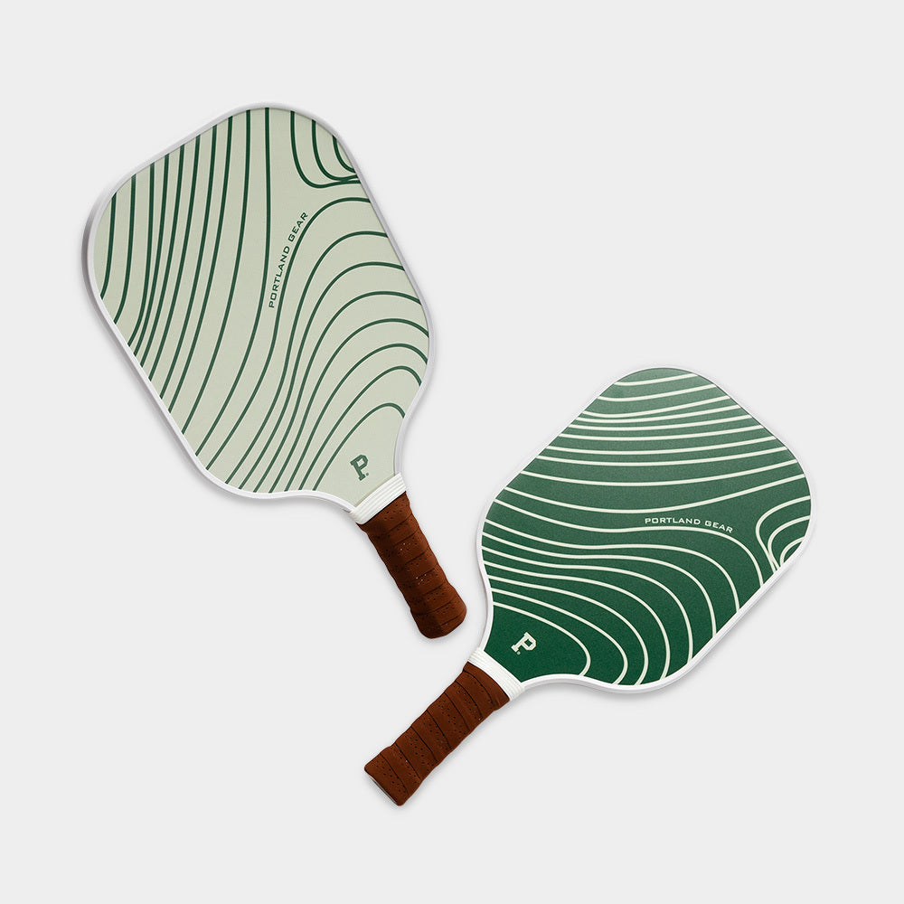 Two Pickleball Paddles from Portland Gear lying on a light grey background. Both paddles feature a white base with dark green curved lines and brown grips. One paddle is slightly lighter in color, while the other is a deeper green. These limited edition paddles exude style and performance.