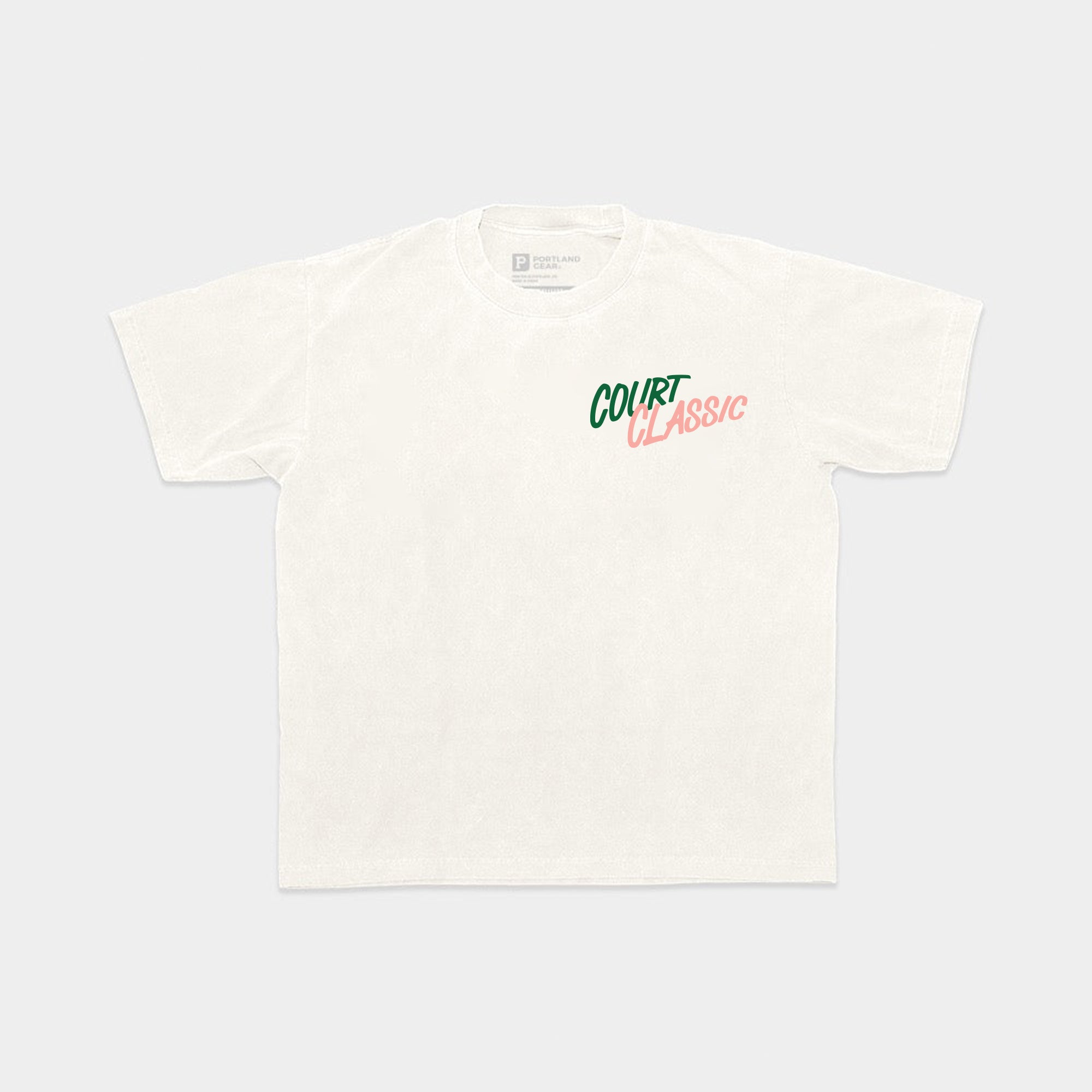 The All-American Court Classic Tee is a white t-shirt crafted from 100% cotton, featuring "COURT CLASSIC" printed on the left chest area. "COURT" is presented in green capital letters, while "CLASSIC" appears in pink with a retro, italicized font. This product comes in sustainable packaging, making it an eco-friendly purchase.