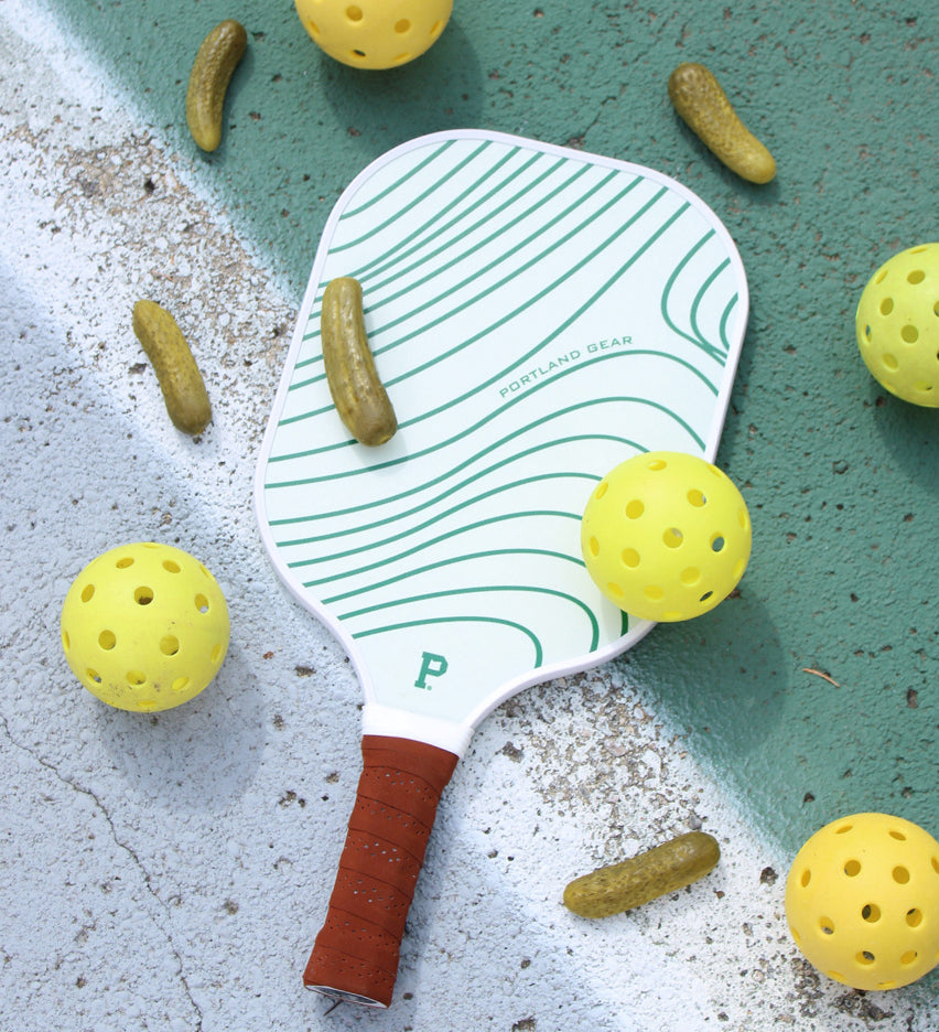 A limited edition Pickleball Paddle with a green and white swirling pattern lies on a green court. Surrounding it are yellow pickleball balls and small pickle-shaped pickles scattered on the surface. The paddle, branded with "Portland Gear," features a leather grip handle.