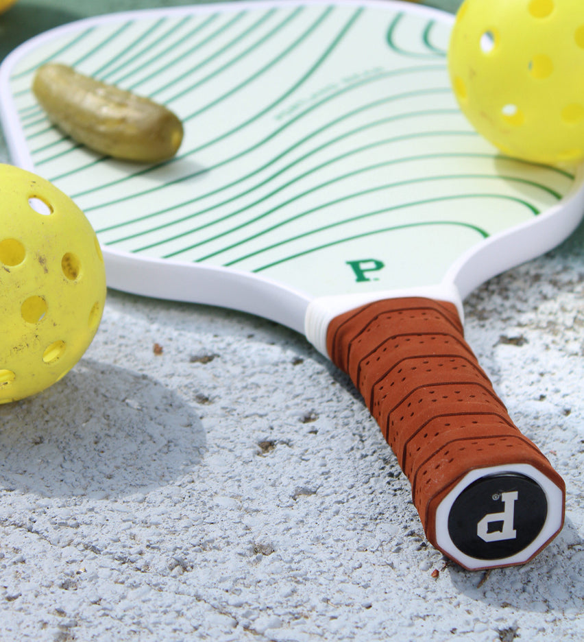 A limited edition Pickleball Paddle in green and white with a brown grip from Portland Gear lies on a concrete surface beside two yellow perforated pickleballs and a pickle.