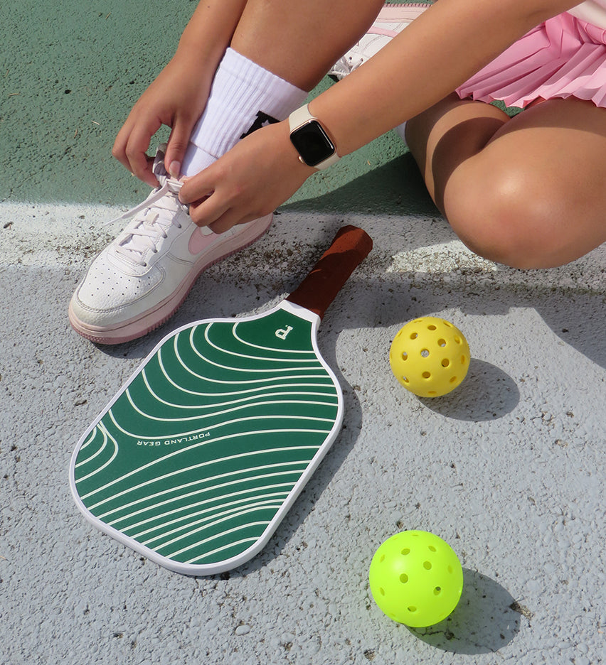 A person tying the laces of their white sneakers while sitting on the ground beside a Pickleball Paddle from Portland Gear and two yellow perforated balls. They are wearing a smartwatch and white socks, with the surface of a tennis court visible in the background.