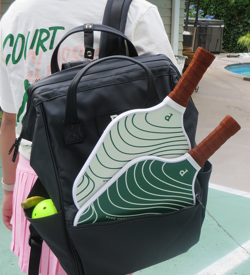 A person wearing a white "Court Classic" t-shirt from Portland Gear and a pink pleated skirt carries a black backpack with two Pickleball Paddles and a yellow pickleball. The scene is outdoors, next to a swimming pool with a green-tiled pool deck.