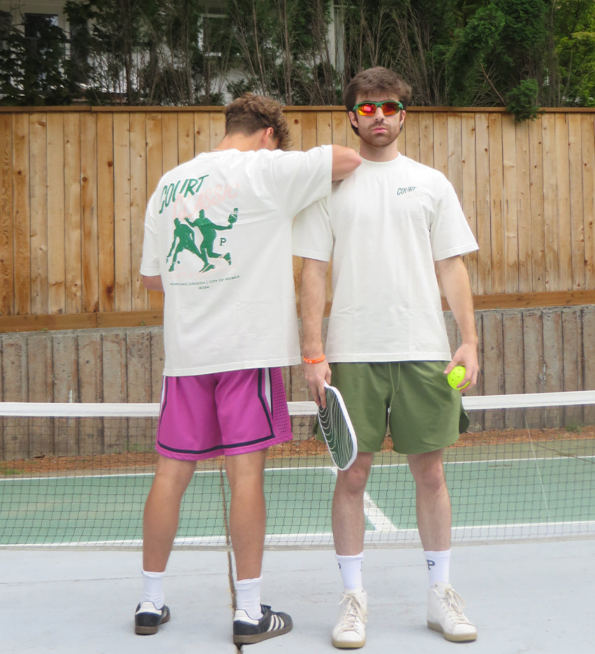 Two men stand on a tennis court. The man on the left, wearing an All-American Court Classic Tee with green graphics and pink shorts, faces away. The man on the right, in 100% cotton attire—white shirt and green shorts—faces forward, holding a tennis ball and racket, ready for their pickleball pack match.